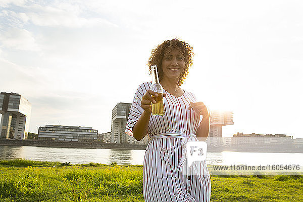Glückliche Frau mit lockigem Haar  die eine Bierflasche hält  während sie am Fluss in der Stadt steht