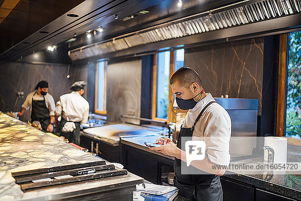 Koch mit Schutzmaske und Smartphone in der Restaurantküche