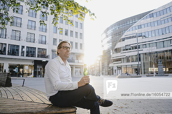 Businessman sitting on a bench in the city at sunset with a bottle of beer