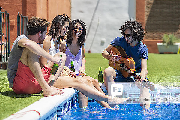 Fröhliche junge Männer und Frauen sitzen mit Gitarre und Bierflaschen am Pool an einem sonnigen Tag