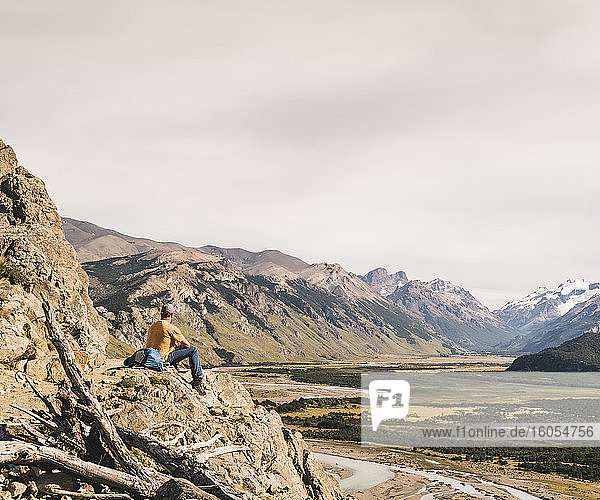 Männlicher Wanderer  der auf einem Felsen sitzend die patagonischen Anden gegen den Himmel betrachtet  Argentinien
