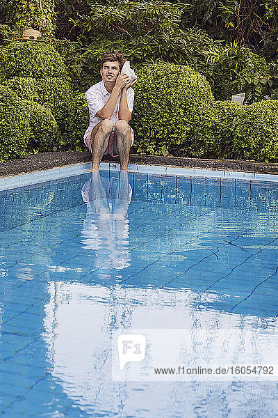 Junger Mann lauscht durch eine Muschel  während er am Pool vor Pflanzen sitzt