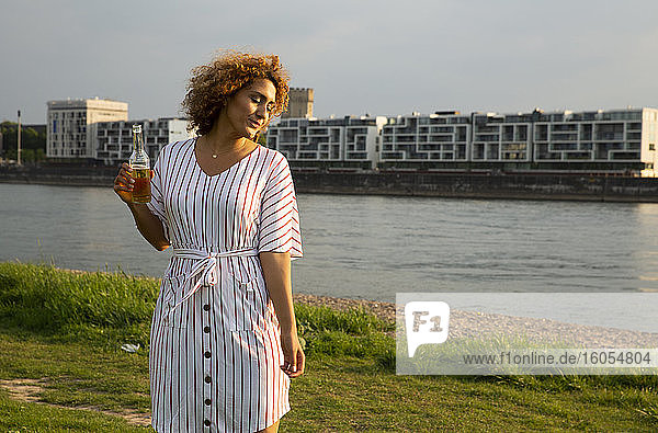 Mid erwachsene Frau hält Bierflasche stehen auf grasbewachsenen Land gegen Fluss in der Stadt bei Sonnenuntergang