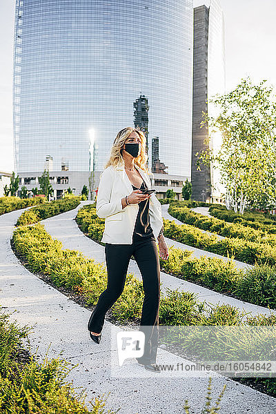 Weiblicher Berufstätiger mit Maske  der ein Smartphone hält  während er auf einem Fußweg in der Stadt spazieren geht