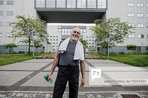 Glücklicher älterer Mann  der eine Wasserflasche hält  während er auf dem Fußweg vor einem Gebäude steht
