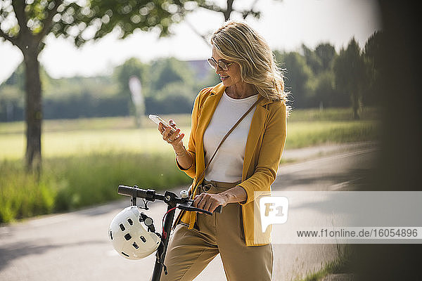 Ältere Frau  die ein Smartphone benutzt  während sie mit einem Motorroller am Straßenrand steht