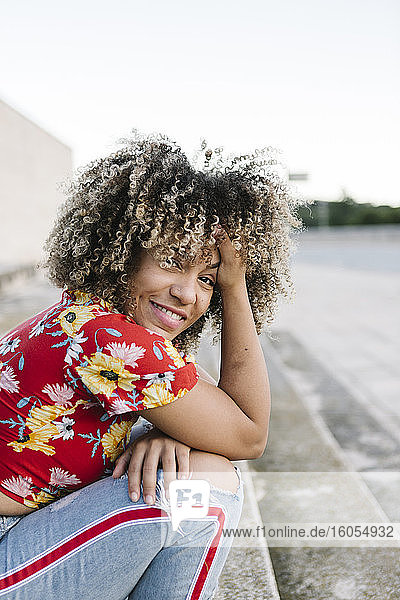 Glückliche junge Frau sitzt mit der Hand im Haar auf einer Treppe an einem sonnigen Tag