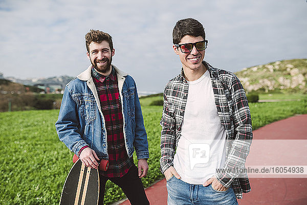 Smiling male friends standing on footpath against cloudy sky in park