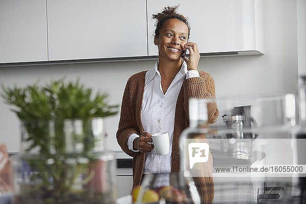 Porträt einer lächelnden Geschäftsfrau am Telefon  die in der Küche steht und mit einer Tasse Kaffee in die Ferne schaut