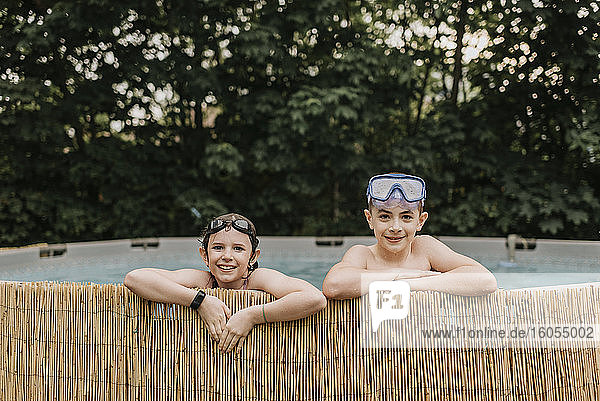 Lächelnde Geschwister mit Schwimmbrillen schwimmen im Pool gegen Bäume im Hof