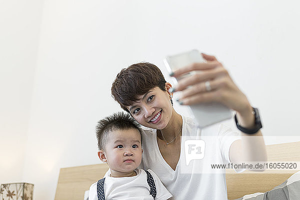 Lächelnde Mutter  die ein Selfie mit ihrem süßen Sohn macht  während sie zu Hause auf dem Bett sitzt