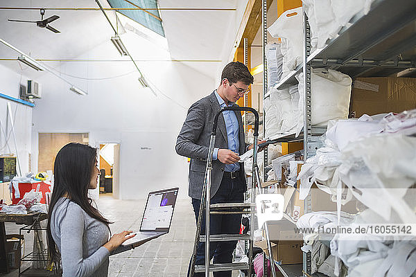 Businessman on step ladder and female employee with laptop in a warehouse