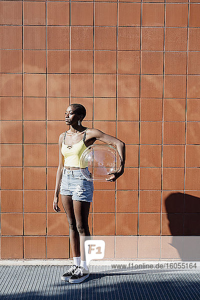 Thoughtful young woman holding glass container while standing on sidewalk in city