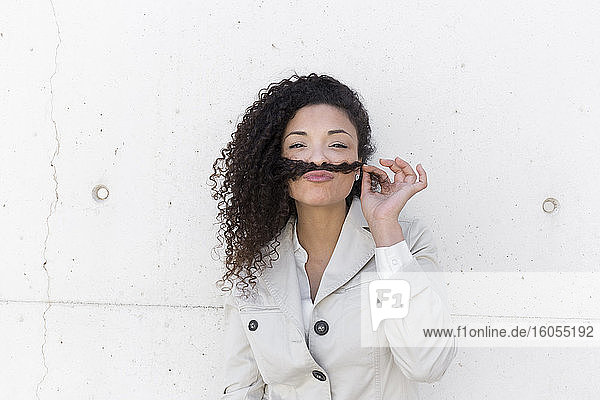 Stylish businesswoman holding hair on lips like mustache against wall