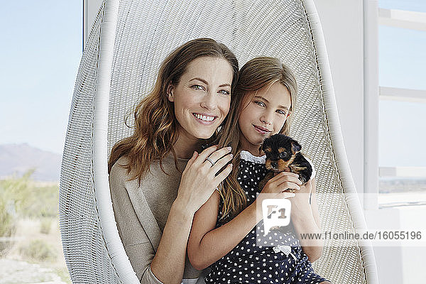 Portrait of smiling mother and daughter sitting in hanging chair with cute puppy