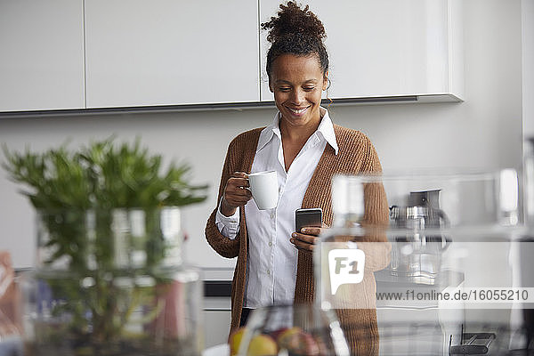 Porträt einer lächelnden Geschäftsfrau  die mit einer Tasse Kaffee in der Küche steht und auf ihr Mobiltelefon schaut