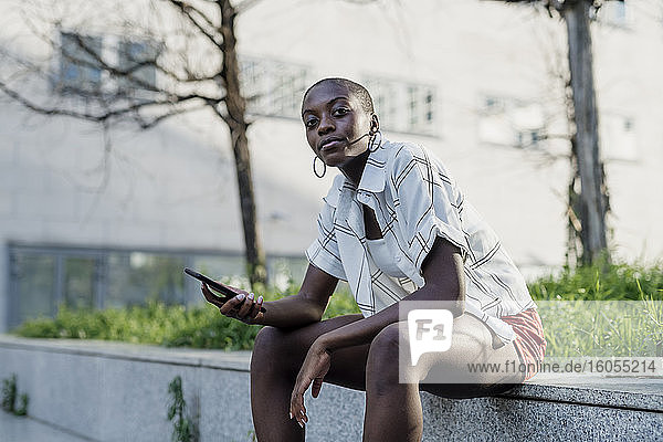 Junge Frau mit rasiertem Kopf  die ihr Smartphone benutzt  während sie auf einem Sitz in der Stadt sitzt