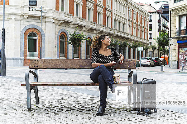 Lächelnde junge Geschäftsfrau in eleganter Freizeitkleidung sitzt auf einer Bank neben einem Gepäckstück