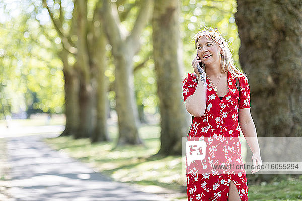 Frau spricht in ihr Smartphone  während sie auf dem Fußweg in einem öffentlichen Park spazieren geht
