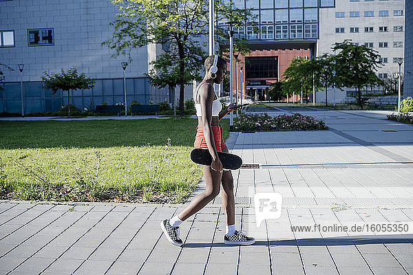 Junge Frau  die ein Skateboard hält und ein Smartphone benutzt  während sie auf einem Fußweg in der Stadt spazieren geht