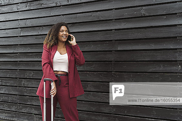 Lächelnde schöne weibliche Unternehmerin  die mit dem Handy spricht  während sie mit Gepäck vor einer schwarzen Holzwand steht