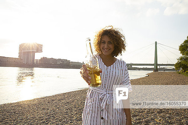 Happy woman with curly hair holding beer bottle while standing on land against river in city