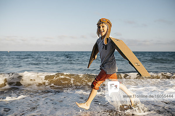 Boy with aircraft wings and cap splashing while running at beach