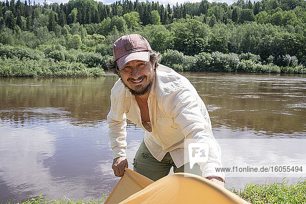 Lächelnder erwachsener Mann  der ein Zelt aufbaut  während er an einem See im Wald steht