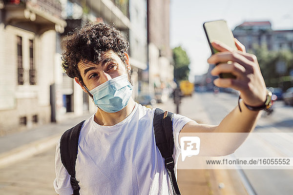 Nahaufnahme eines jungen Mannes mit Maske  der ein Selfie mit seinem Smartphone macht  während er in der Stadt steht