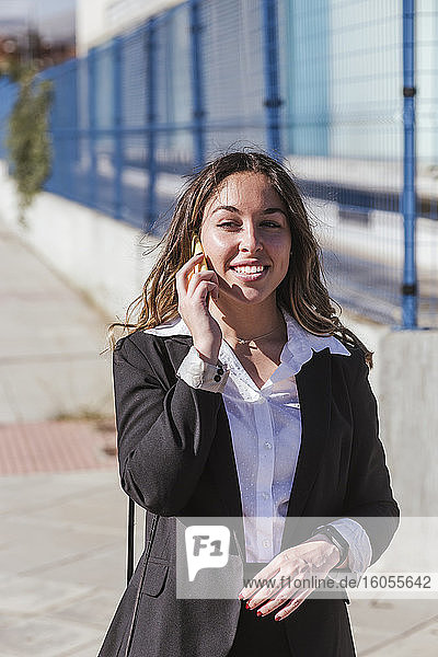 Lächelnde Geschäftsfrau  die über ein Smartphone spricht  während sie an einem sonnigen Tag in der Stadt steht
