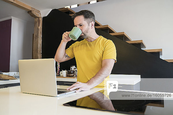 Mann trinkt Kaffee und benutzt einen Laptop auf einer Kücheninsel zu Hause