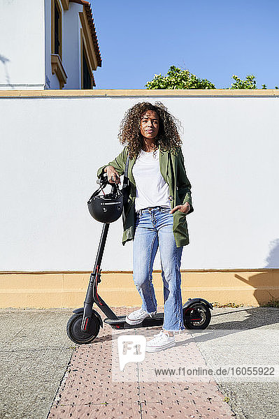 Confident woman standing with electric push scooter on sidewalk during sunny day