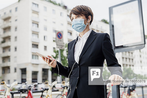 Geschäftsfrau mit Maske  die ein Smartphone hält  während sie in der Stadt steht