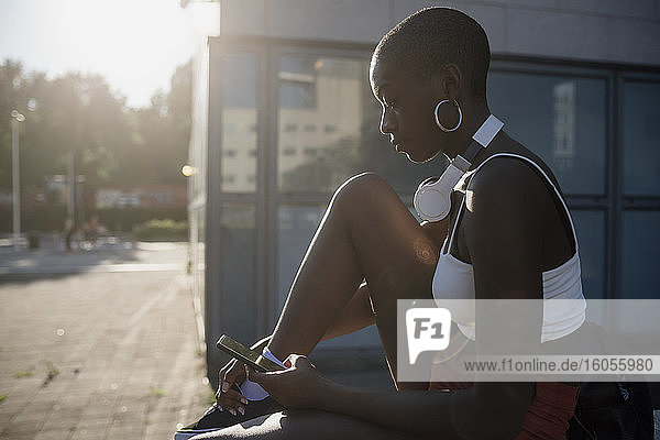 Junge Frau mit rasiertem Kopf  die ein Smartphone benutzt  während sie an einem sonnigen Tag in der Stadt sitzt