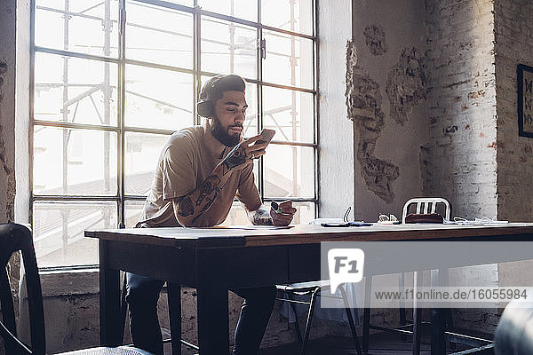 Creative young businessman with headphones and smartphone sitting at table in loft office