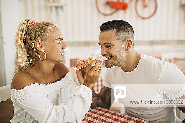 Cheerful young woman feeding pizza to boyfriend while sitting in restaurant