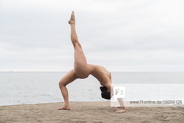 Biegsame junge nackte Frau übt das Rückwärtsbeugen am Strand gegen den Himmel