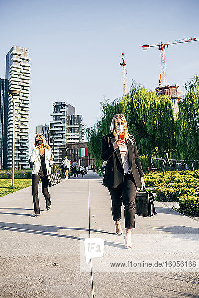 Weibliche Berufstätige  die Masken tragen und ein Smartphone benutzen  während sie auf einem Fußweg in der Stadt gehen
