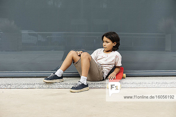 Thoughtful boy with skateboard relaxing on footpath against wall