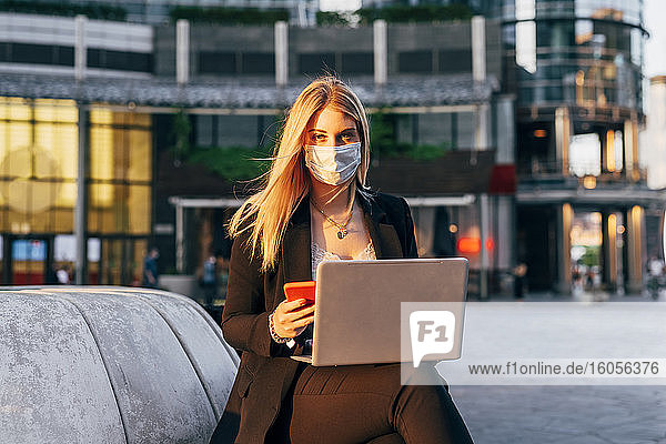 Businesswoman wearing mask using laptop and smart phone while sitting against building