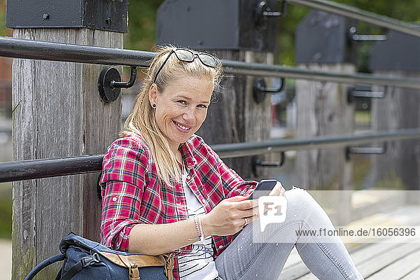 Lächelnde Frau im mittleren Erwachsenenalter  die ein Smartphone benutzt  während sie an der Reling auf dem Pier sitzt