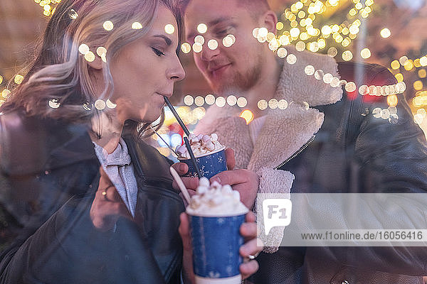 Man sharing his hot chocolate with girlfriend in store at amusement park seen through window