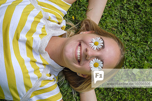 Nahaufnahme einer lächelnden Frau mit Gänseblümchen auf den Augen  die auf einer Wiese im Park liegt