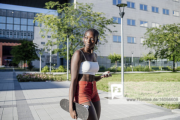 Junge Frau hält Handy und Skateboard und schaut weg  während sie auf einem Fußweg in der Stadt steht