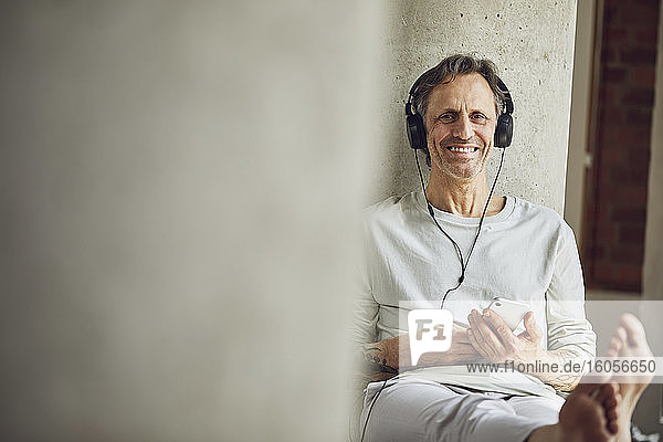 Porträt eines lächelnden älteren Mannes mit Kopfhörern  der in einer Loftwohnung Musik hört