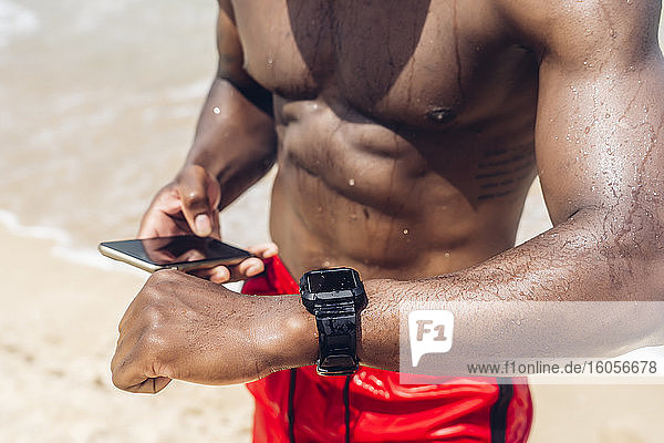 Sportlicher Mann mit Smartphone und Smartwatch am Strand