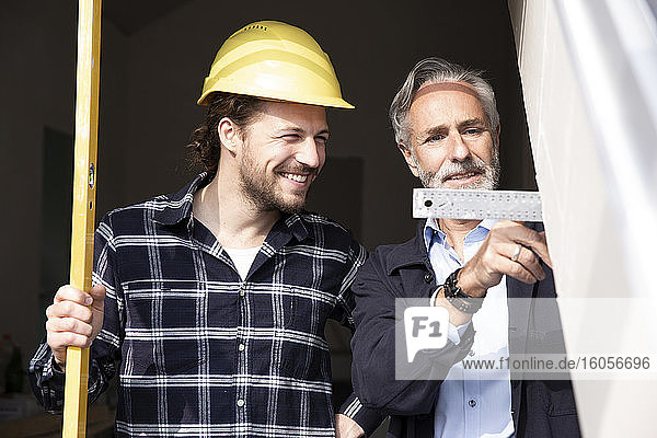 Smiling construction worker looking at measurement showing by architect in constructing house seen through window
