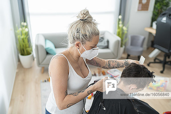 Friseurin mit Maske schneidet einem Jungen zu Hause während der Ausgangssperre die Haare mit einem elektrischen Rasiermesser