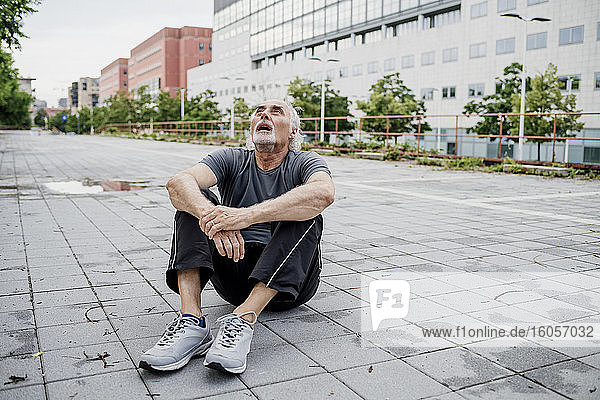Müder älterer Mann mit offenem Mund  der nach oben schaut  während er auf einem Fußweg in der Stadt sitzt