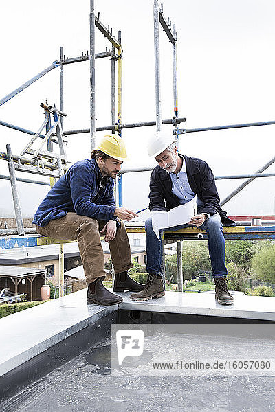 Architect and construction worker discussing blueprint while sitting on scaffold against clear sky